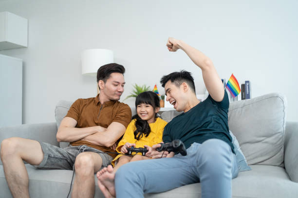 asian male gay family with young daughter play game together in house. little child daughter sit on sofa with attractive romantic male lgbt couple play joystick game at home. homosexual-lgbtq concept. - gamer watching tv adult couple imagens e fotografias de stock