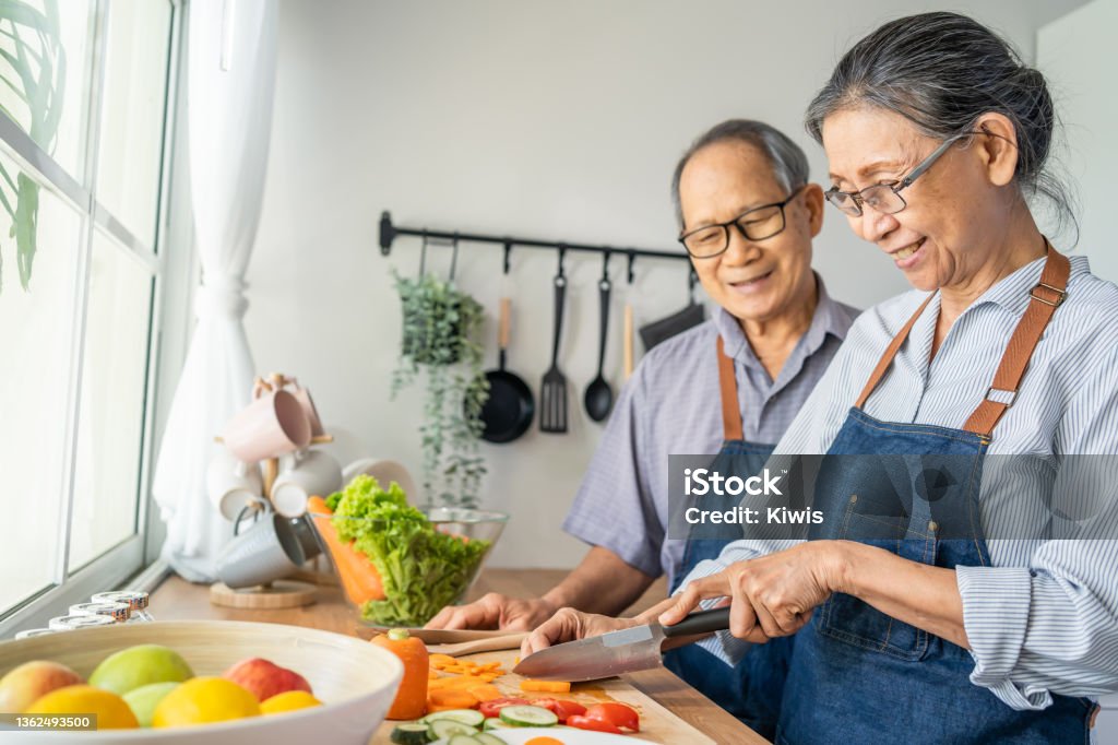 Asian loving senior elderly couple wear apron and cooking in kitchen. Attractive strong old man and woman grandparent wear eyeglass enjoy retirement life activity at home. Family relationship concept. Senior Adult Stock Photo