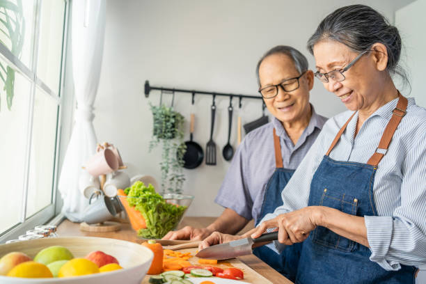 Asian loving senior elderly couple wear apron and cooking in kitchen. Attractive strong old man and woman grandparent wear eyeglass enjoy retirement life activity at home. Family relationship concept. stock photo