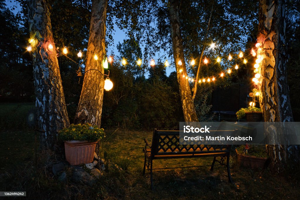 string of lights in the garden with garden bench and sheltered corner hidden by hedges in the evening. string of lights in the garden with garden bench and sheltered corner hidden by hedges in the evening. romantic weekend time, to celebrate or relax. Yard - Grounds Stock Photo