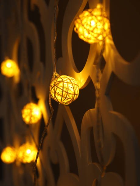 istock Yellow Rattan ball garland on patterned wall. Christmas led lights on dark background. Blurred glowing light bulb garland. Closeup, vertical photo. 1362496248