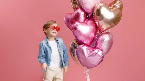 Photo of Boy dancing, smiling and having fun near  big branch of pink heart-shaped balloons. Isolated on pink background.