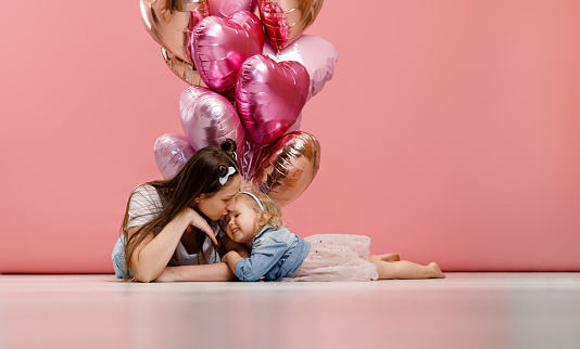 Young mother and her daughter dancing, smiling and having fun near  big branch of pink heart-shaped balloons. Isolated on pink background.