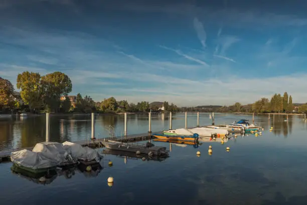 Boats lie on a wooden jetty on the Rhine near the village of Stein am Rhein. Trees and houses on the shore, blue sky with clouds.