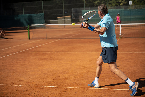 Active senior couple playing tennis on a clay court during sunny day.