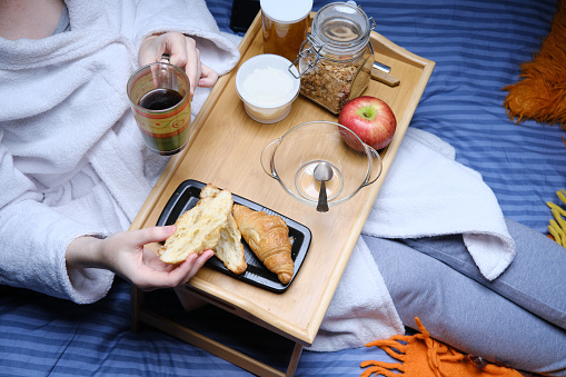 Mature woman wearing bathrobe and pajamas is sitting at her bed and having breakfast