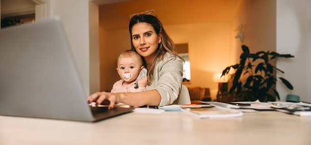 Single mom typing on a laptop while working in her home office. Multitasking mom working on a new creative project at her desk. Female interior designer carrying her baby on her laps.