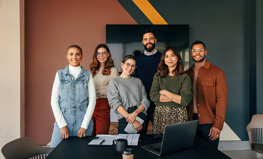 Successful business team smiling at the camera while standing behind a table in a boardroom. Group of multicultural businesspeople working together in a modern workplace.