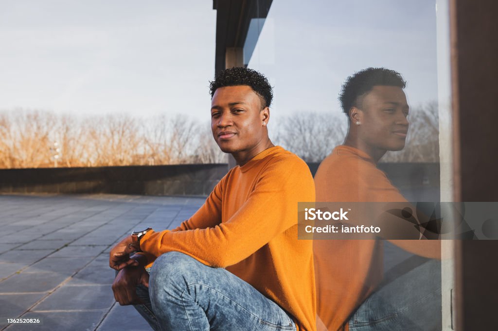 Young afro-american man wearing orange sweater reflected in a glass surface. Young afro-american man wearing orange sweater reflected in a glass surface. City life concept. Adult Stock Photo