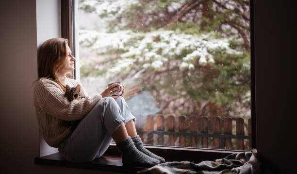 giovane ragazza allegra seduta a casa sul davanzale della finestra in un maglione caldo che beve caffè con il gattino grigio in braccio - snow heat adult blond hair foto e immagini stock