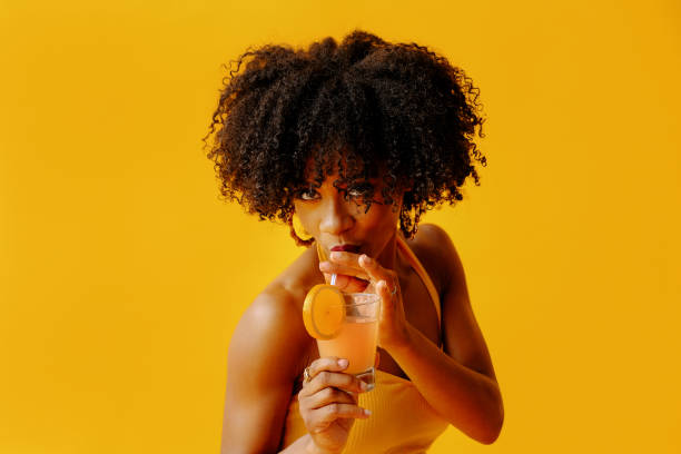 belle jeune femme afro-américaine en tenue d’été lumineuse buvant cocktail rafraîchissant isolé sur fond jaune - fruit women beautiful food photos et images de collection