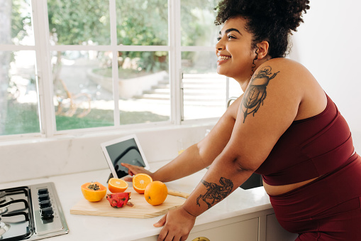 Happy woman standing with fruits and tablet pc at kitchen. Plus size female in fitness wear making healthy food in kitchen.