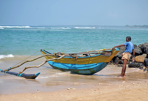 Lone fisherman and outrigger canoe waits by rocky outcrop on Hikkaduwa Beach, Sri Lanka. South Asia. Sri Lanka, formerly Ceylon, is a  beautiful warm tropical island off the south eastern coast of India with a rich history of Sinhalese kingdoms and a mixture of faiths including Hindu Buddhist and Tamil as well as Dutch and British colonial influences. It has a wealth of birdlife and wildlife including many endangered wild animals.