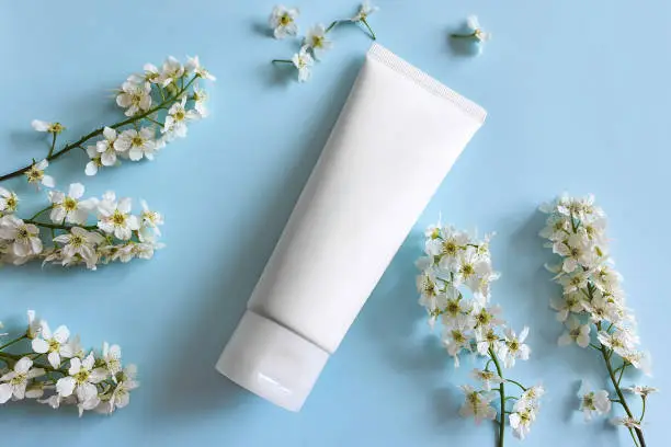 Photo of White squeeze bottle plastic tube for branding of medicine or cosmetics - cream, gel, skincare, moisturize. Cosmetic container and white bird cherry flowers on blue background. Top view, mockup.