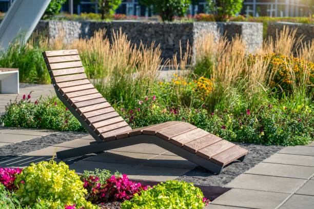 empty brown wooden deck chair or chaise longue on tile among decorative grass and flowers in recreation area. garden landscape with chairs in city park. concept of recreation, tanning in yard - tanning bed brown relaxation resting imagens e fotografias de stock