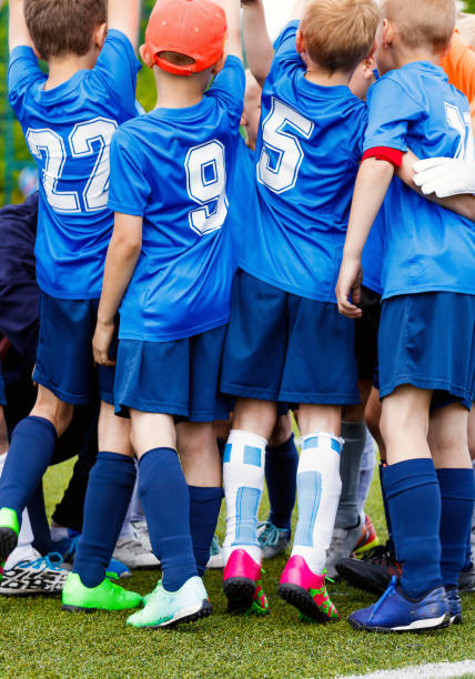 vertikales bild von glücklichen jungs in der sportmannschaft, die gemeinsam erfolge feiern. schulkinder in blauen sportlichen trikothemden mit weißen nummern. kinder treiben sport auf rasenplatz - mannschaftsfußball stock-fotos und bilder