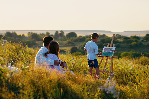 Family spending time together in nature. Parents sitting in meadow, their little son standing nearby and painting on canvas. Creative hobby. Concept of arts