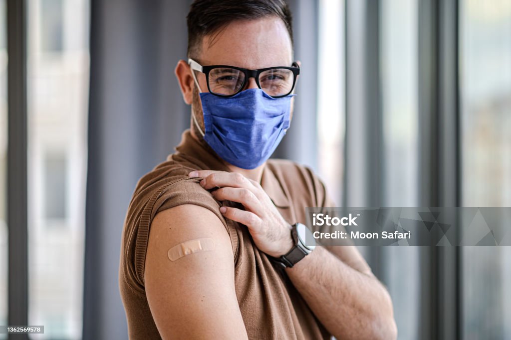 Young adult man showing pointing to bandage on his arm shoulder wearing protective mask done with vaccination. Male just got vaccinated against Covid19 face mask and eyeglasses looking at camera. Booster Dose Stock Photo