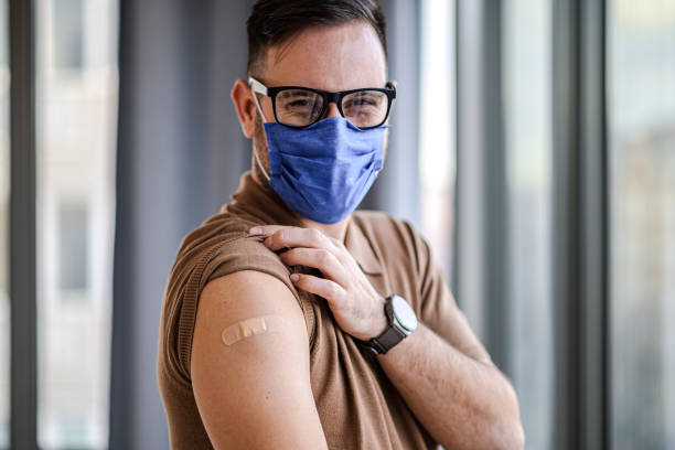 Young adult man showing pointing to bandage on his arm shoulder wearing protective mask done with vaccination. Male just got vaccinated against Covid19 face mask and eyeglasses looking at camera. Young adult man showing pointing to bandage on his arm shoulder wearing protective mask done with vaccination. Male just got vaccinated against Covid19 face mask and eyeglasses looking at camera. booster dose photos stock pictures, royalty-free photos & images