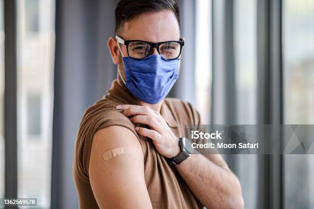 Young Adult Man Showing Pointing To Bandage On His Arm Shoulder Wearing Protective Mask Done With Vaccination Male Just Got Vaccinated Against Covid19 Face Mask And Eyeglasses Looking At Camera Stock Photo - Download Image Now