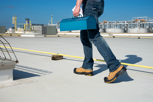 A mechanic or tradesperson carrying a toolbox on the roof of an industrial building.