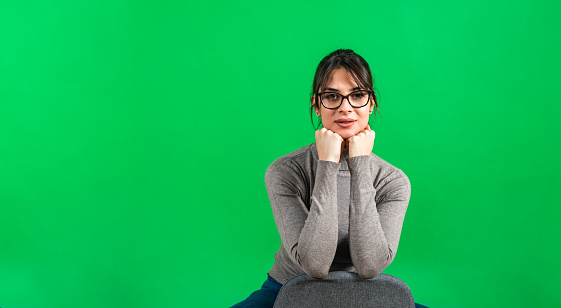 Education, school and people concept - happy smiling young pretty student woman or teacher in glasses over green chalkboard background sitting on the chair holding head in her hands, not isolated