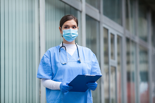 Female NHS doctor standing outside of medical healthcare facility,wearing blue uniform,protective surgical face mask,blue gloves and stethoscope,holding patient clipboard form,looking away pensive