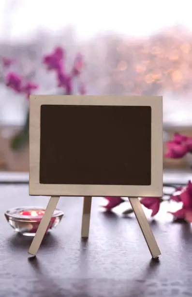 Greeting card mockup, copy-space on blackboard, chalk board. Small decorative black board with pink flowers, orchid and magnolia. Aromatic candle, tea light. Sunset light out of the window.