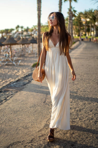 bella donna bruna che cammina sulla spiaggia del tramonto in maxi abito alla moda, rilassandosi nelle lussuose vacanze estive tropicali. - vestito da donna foto e immagini stock