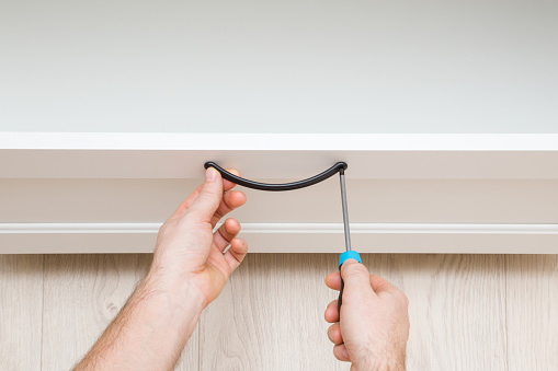 Young adult man hand using manual screwdriver and screwing black metal drawer handle. Assembling new white wooden furniture. Closeup. Point of view shot.