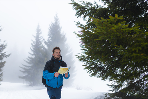 A young male hiker, wearing a blue jacket, is lost in a foggy mountain. He is looking at the map and his smart phone to try and find a route.