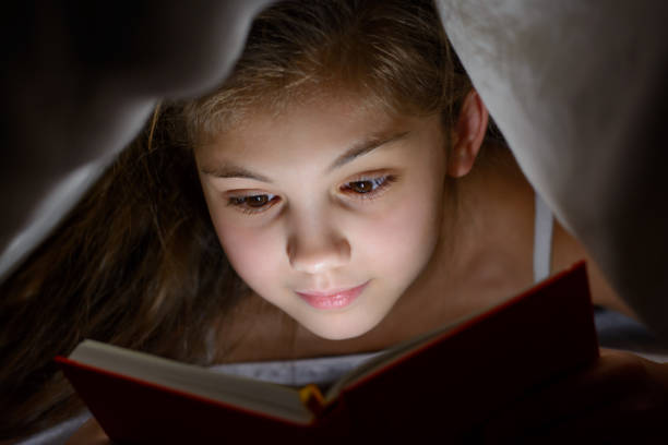 istock Small girl reads book with a flashlight under the blanket in bed 1362603125