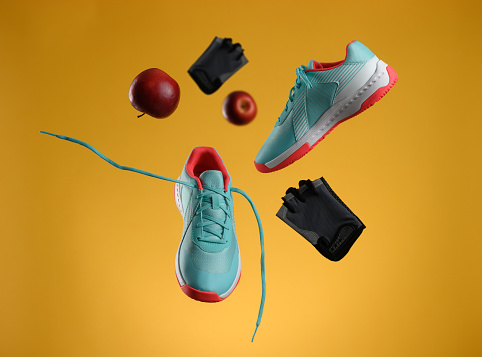 red apple, gloves and pair of textile blue sneakers with laces levitate on a yellow background. Shoes for sports, jogging