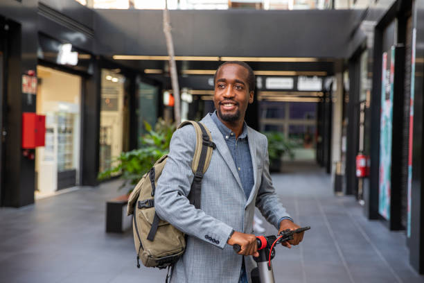 uomo d'affari africano con il suo scooter elettrico. ragazzo sorridente elegantemente vestito va a lavorare con un veicolo non inquinante - concetto di mobilità pulita - nonpolluting foto e immagini stock