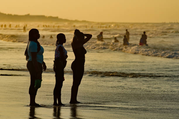 Dakar. Senegal. October 13, 2021. Three city girls standing in the seawater watching the locals bathing in the Atlantic Ocean flooded with sunset sunlight.