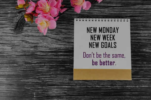 Positive business motivational message on calendar cover page - New Monday. New Week. New Goals. Don't be the same. Be better. With pink flower decoration on rustic wooden table background.