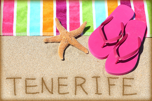 Tenerife beach travel concept background. TENERIFE written in sand with water next to beach towel, summer sandals and starfish. Summer and sun vacation holidays on Canary Islands, Spain.