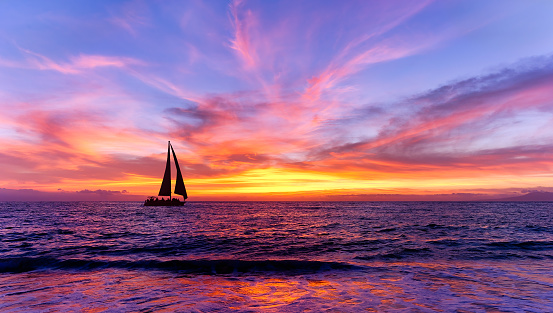 A Sailboat Is Sailing Along The Ocean With a Flock Of Birds Flying Overhead Against A Colorful Sunset Sky