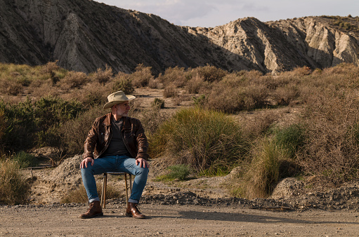 Adult man in cowboy hat sitting on abandoned chair in Tabernas desert, Almeria, Spain