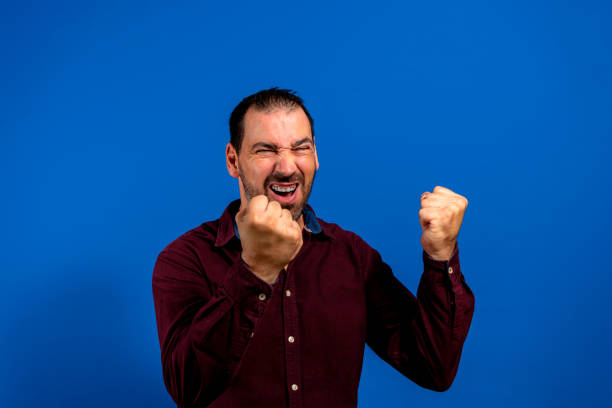 istock Hispanic man with beard celebrating energetically with clenched fists, isolated on blue studio background. 1362668759