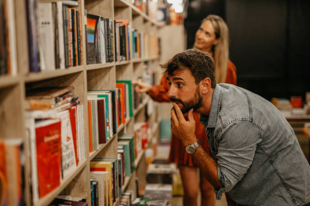Spontaneous image of a cute bearded young adult male looking for a book to buy, going through the books and reading while next to him is a beautiful blonde woman, doing the same. Both elegant casually dressed, loving their favorite book store place