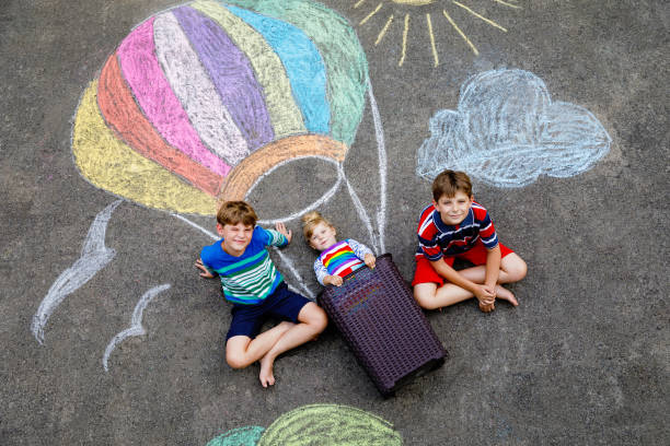 幸せな小さな幼児の女の子と熱気球で飛ぶ2人の子供の男の子は、夏に地面やアスファルトに虹色の色でカラフルなチョークで描かれました。3人の子供、楽しい兄弟 - child balloon outdoors little boys ストックフォトと画像
