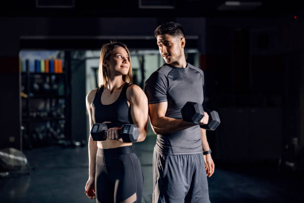 un couple sportif heureux faisant des exercices pour les biceps avec des haltères dans un gymnase et établissant un contact visuel les uns avec les autres. - gym muscular build weights two people photos et images de collection