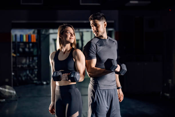 A happy sporty couple doing exercises for biceps with barbells in a gym and making eye contact with each other. A happy sporty couple doing exercises for biceps with barbells in a gym and making eye contact with each other. images of female bodybuilders stock pictures, royalty-free photos & images