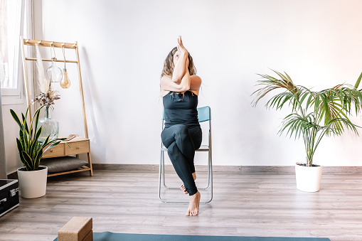 Full body barefoot female in black activewear twisting arms and legs while sitting on chair and doing Garudasana pose during yoga session in light modern studio