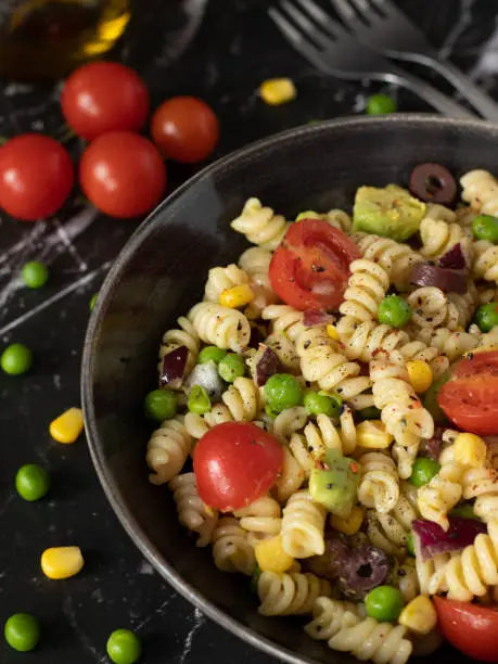 Vegan colorful Italian pasta salad in a bowl with cherry tomatoes, green peas, corn, avocado, olives, olive oil, and spices. A close-up of healthy plant-based macaroni with fresh vegetables for dinner