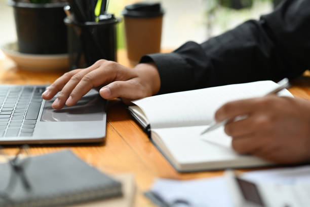 istock Cropped image of a young office man writing on a notebook while using a computer laptop at the modern working desk. 1362695822
