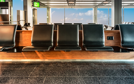 Horizontal row of empty leather bench chairs, modern design with wood panel in front of window frame with blue sky. Waiting area of airport lounge.