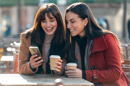 Shot of two attractive friends enjoying coffee together while looking smartphone and talking sitting on the terrace of a bar.
