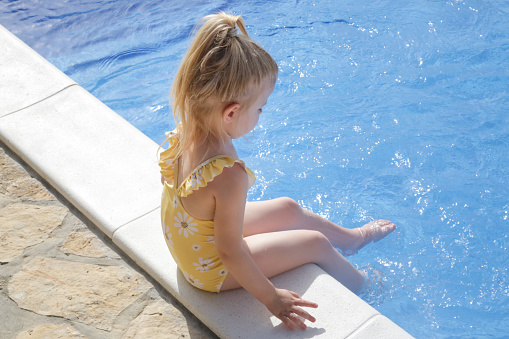 Toddler girl in yellow swimsuit sitting at the edge of pool